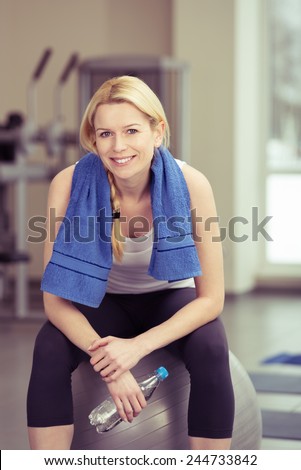 Young woman taking a break from her exercises to drink a bottle of water in a gym sitting on a pilates ball smiling at the camera