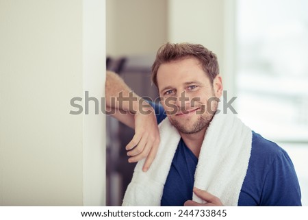 Fit young man in a gym standing with a towel around his shoulders in front of equipment smiling at the camera in a healthy lifestyle concept
