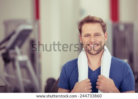 Handsome young man posing in a gym with a towel around his shoulders looking at the camera with a beaming smile full of vitality