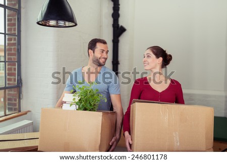 Young couple moving into a new house standing side by side holding packing cartons looking at each other with a smile