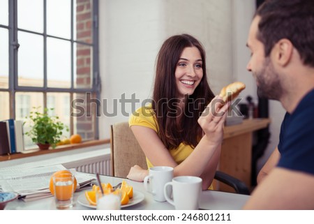 Loving wife feeding her husband his breakfast with a playful smile as she holds out a roll for him to bite
