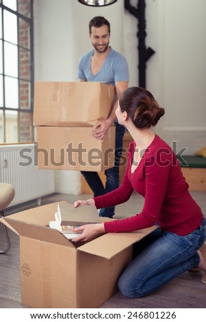 Happy young couple moving into a new home smiling at each other as the wife unpacks a carton on the floor as the husband arrives with two more
