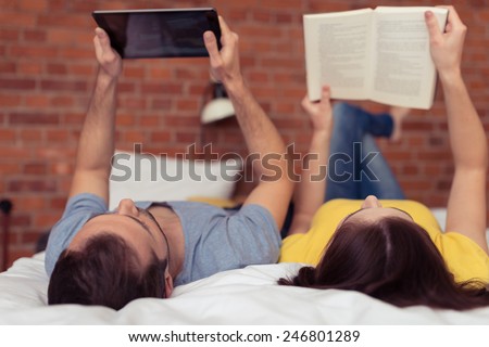 Young couple lying on their backs reading on the bed , the husband using a tablet and the wife a book