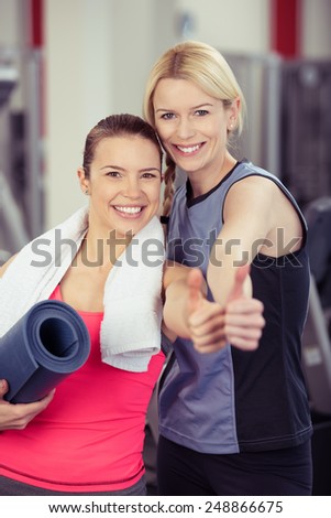 Two happy young women standing close together in a gym giving a thumbs up with rolled exercise mats under their arms to show their approval of a healthy lifestyle