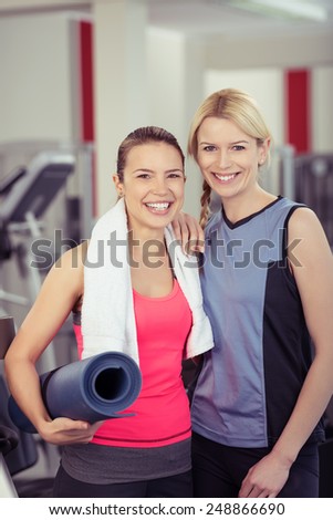 Two attractive young fit female friends at the gym standing side by side with a rolled up exercise mat smiling happily at th camera