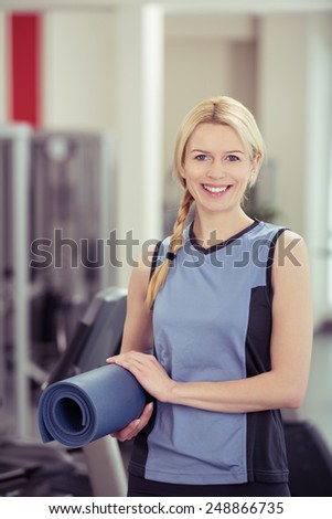 Pretty blond woman carrying a rolled up exercise mat as she walks past the equipment in the gym looking at the camera with a smile