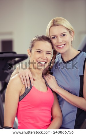 Two attractive happy healthy fit young women posing arm in arm at the gym smiling at the camera in a health and fitness concept