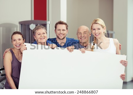 Group of Healthy People Smiling Behind Empty White Card Board While at Inside the Fitness Gym. Emphasizing Copy Space.