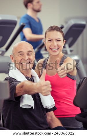 Smiling older man and young woman in a gym standing together amongst the equipment during a workout giving an enthusiastic thumbs up gesture of approval