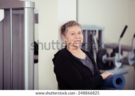 Smiling Senior Woman Holding Workout Mat While Leaning on the Post at the Fitness Gym