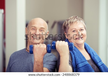 Fit elderly couple working out with weights each holding a dumbbell in a flexed hand as they tone their muscles and looking at the camera with happy smiles