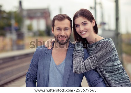 Happy couple waiting at a railway station standing on the platform in a close embrace smiling at the camera