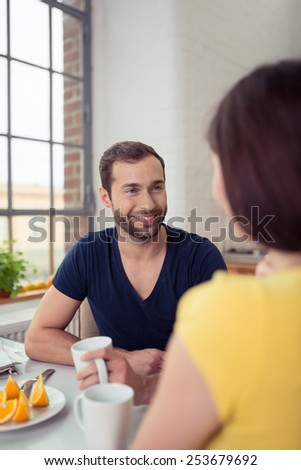 Smiling bearded man enjoying breakfast smiling at his wife as he drinks a mug of coffee while seated at the table, over the shoulder view