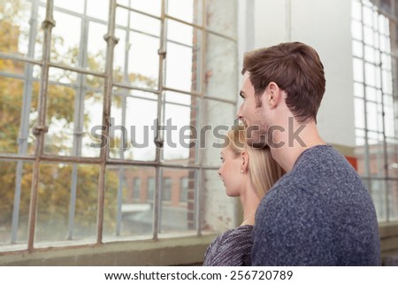 Young couple standing close together in an affectionate embrace lost in thought staring out of a large window in their apartment at an urban street