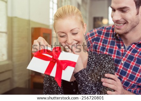 Young man surprising his sweetheart with a gift tied in a large red bow for Valentines Day as he approaches her from behind in an intimate embrace