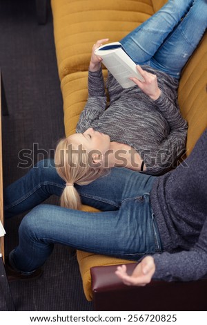 Young woman relaxing with a book at home lying back on the sofa with her head resting on her husbands leg, view from above