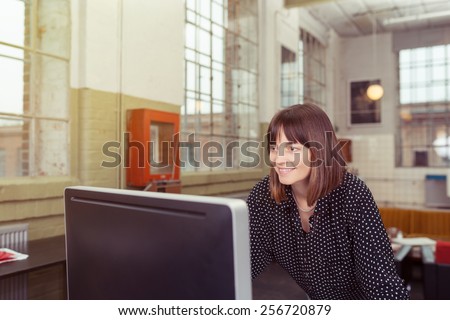 Smiling businesswoman working alone at a large screen desktop computer in a spacious office with a smile on her face