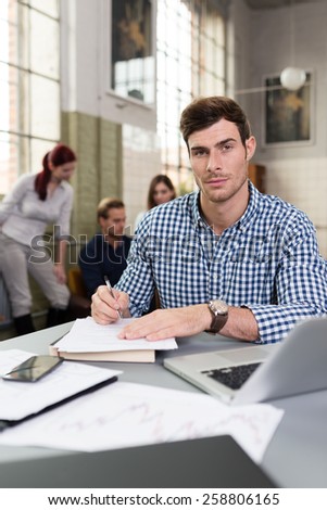 Young businessman working in the office sitting at his desk doing paperwork with colleagues working in the background