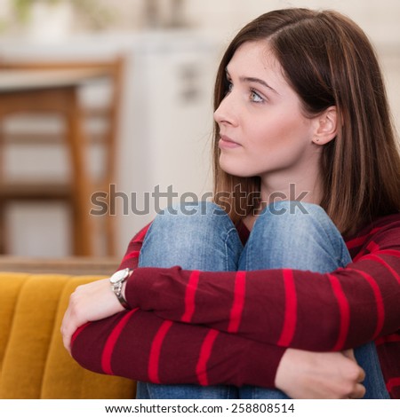 Pretty young brunette woman sitting thinking hugging her raised knees with her arms as she stares off to the side
