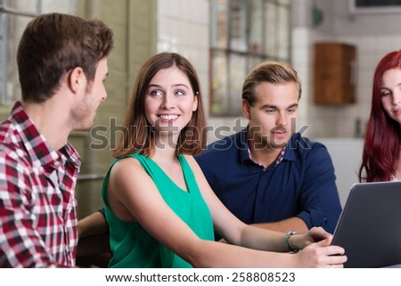 Young man and woman smiling and feeling attracted to each other while sitting at a table next to their young colleagues, in front of a laptop, indoor
