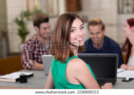 Smiling confident young businesswoman working in a busy office turning to look at the camera