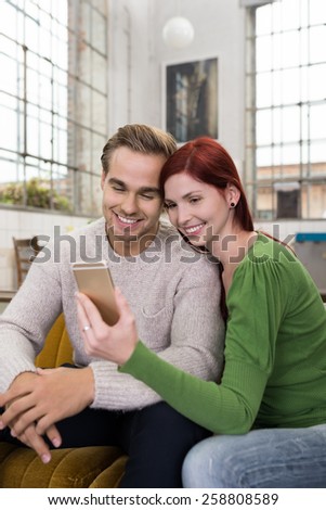 Sweet Smiling White Couple in Casual Outfit, Sitting on the Couch at the Living Room While Watching Something at Mobile Phone.