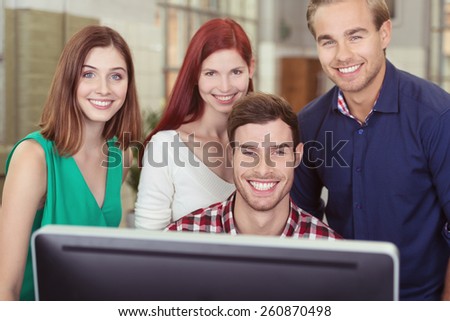 Happy successful young business team standing grouped around a desktop monitor looking at the camera with beaming motivated smiles of satisfaction