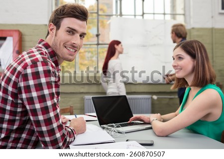 Smiling handsome man at work in the office sitting at a table talking to an attractive young female colleague