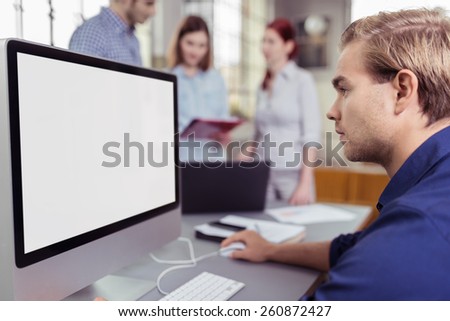 Young businessman working at a large screen computer with a blank white screen visible to the viewer with colleagues chat in the background