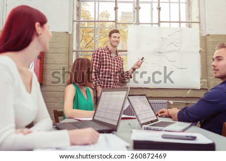 Handsome smiling team leader giving a presentation to his team pointing to a hand drawn graph as they sit around a table watching and following on their laptops