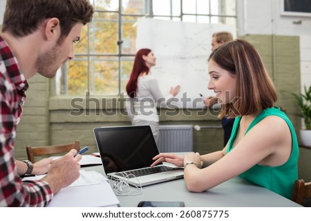 Young man and woman working in an open-plan modern office sitting at a table over a laptop having a discussion as colleagues work in the background