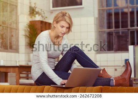 Pretty Blond Woman Surfing the Internet Using her Laptop Computer While Sitting on the Table Behind the Sofa at the Living Room.