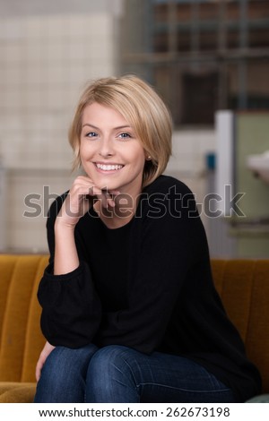 Close up Pretty Smiling Blond Woman in Casual Outfit, Sitting on the Couch with Hand on the Chin While Looking at the Camera.