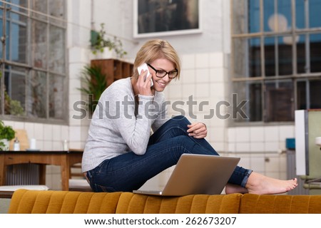 Young woman working from home sitting barefoot on a kitchen counter chatting on her mobile while reading the screen of her laptop balanced on the back of the sofa