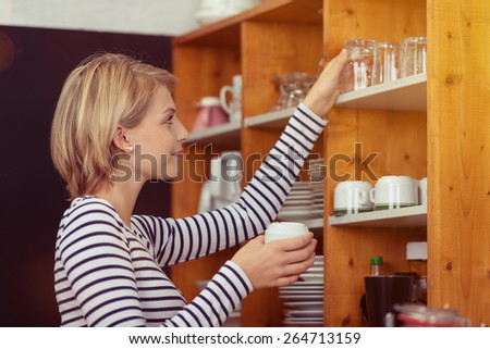 Side View of a Pretty Young Woman with Short Blond Hair Arranging the Glasses at the Wooden Cabinet
