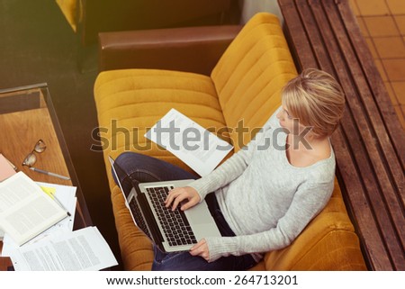 View from above of a young woman sitting cross legged on a sofa studying online on her laptop at home surrounded by her notes