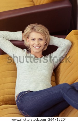 Close up Happy Young Blond Woman Resting on Sofa at the Living Area with Both Hands Behind her Head While Smiling at the Camera.