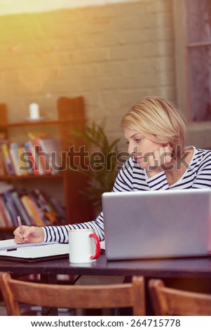 Serious Young Blond Woman Writing on a Paper at the Table, with a Cup of Coffee and Laptop Computer on the Side, While Leaning on her Hand