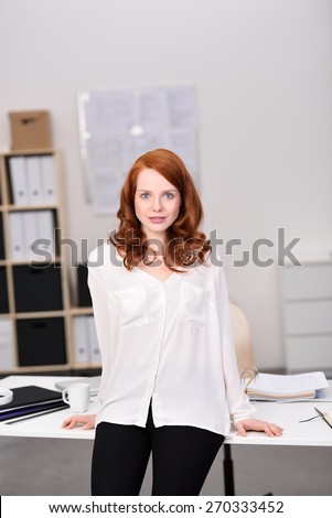 Pretty Young Woman in Black and White Outfit Leaning Back Against the Desk Inside the Office While Looking at the Camera.