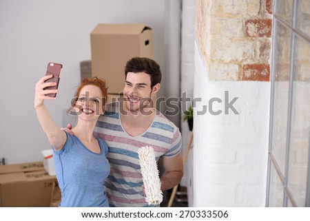 Happy couple posing for a selfie in their new home standing close together with a paint roller alongside an unfinished white wall Happy couple posing for a selfie in their new home standing close together with a paint roller alongside an unfinished white wall