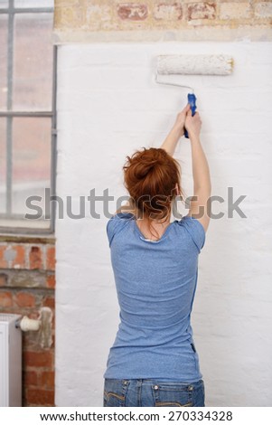Young woman standing with her back to the camera painting a white wall with a roller in a DIY and renovation concept