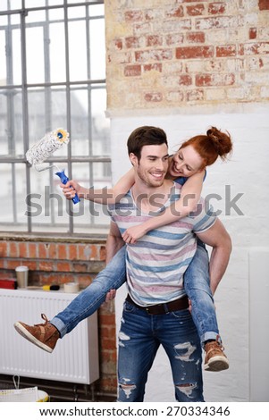 Mischievous young woman riding piggy back on her husbands back as she brandishes a paint brush full of white paint while renovating the apartment