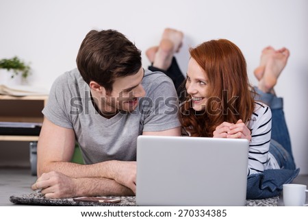 Barefoot young couple relaxing with their laptop lying side by side on the floor at home smiling at each other