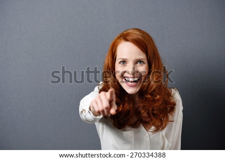 Playful pretty young redhead woman pointing at the camera and laughing with a beaming smile, over grey with copyspace