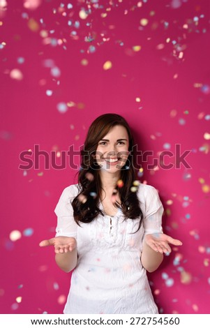Smiling Pretty Girl in White Shirt Opening her Both Hands on a Shower of Confetti, Isolated on Pink Background.