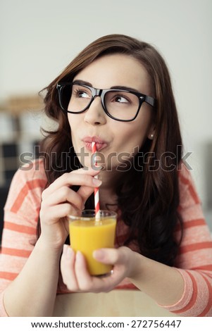Thoughtful young woman in heavy rimmed glasses sitting sipping orange juice and looking off to the side