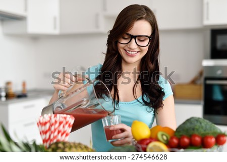Happy healthy young woman wearing glasses pouring vegetable smoothies freshly made from assorted vegetable ingredients on her kitchen counter