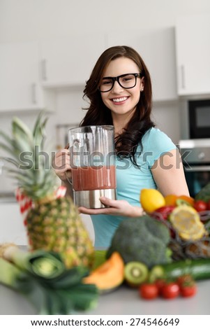 Happy healthy woman making smoothies from a variety of fresh fruit and vegetables on her kitchen counter standing holding a blender with fresh prepared beverage as she smiles at the camera