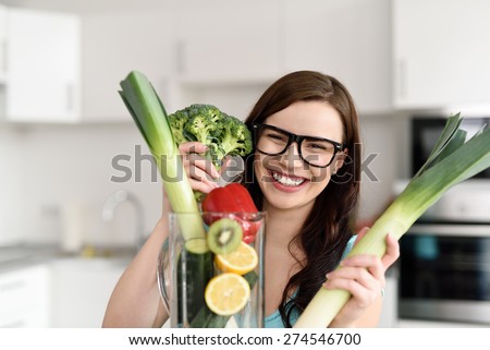 Happy Pretty Girl Holding Fresh Veggies Behind a KItchen Blender Device, Filled with Sliced Fruit and Veggies, and Smiling at the Camera