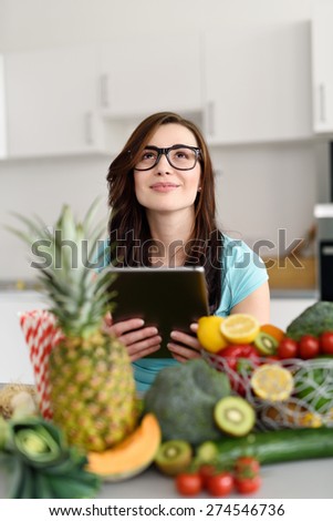 Smiling Thoughtful Girl Looking Up, Holding Tablet Computer at the Kitchen Behind Fresh Fruits and Veggies.